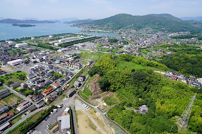 空中写真（北から）中央の山が沖の城跡・手前の空き地は水除浜塩田跡<br />写真左側の宅地は昭和の埋め立て